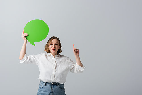 happy overweight woman in white shirt holding green speech bubble and pointing with finger isolated on grey