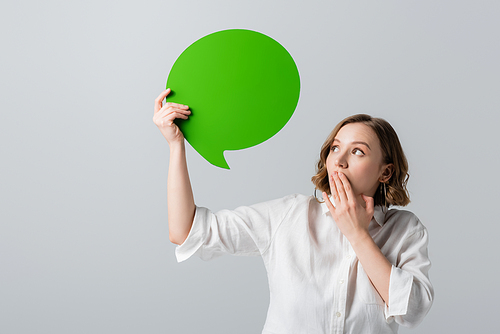 overweight woman in white shirt holding green speech bubble and covering mouth isolated on grey