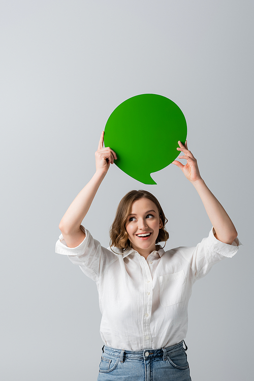 cheerful young woman in white shirt holding green speech bubble above head isolated on grey