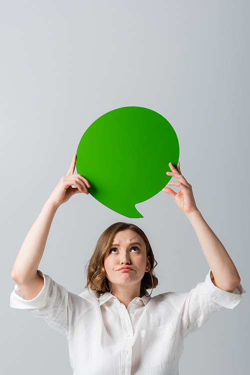 displeased overweight woman in white shirt holding green speech bubble above head isolated on grey