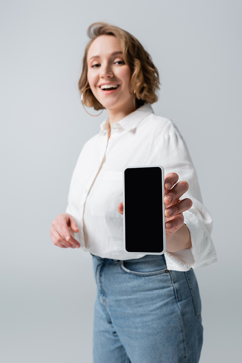 blurred overweight woman holding smartphone with blank screen and smiling isolated on grey