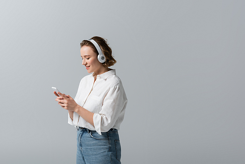 smiling plus size woman in wireless headphones listening music and holding smartphone isolated on grey