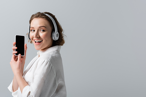 happy plus size woman in wireless headphones listening music and holding smartphone with blank screen isolated on grey