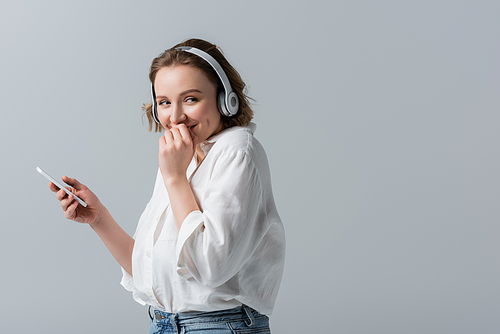 happy plus size woman in wireless headphones listening music and holding smartphone isolated on grey