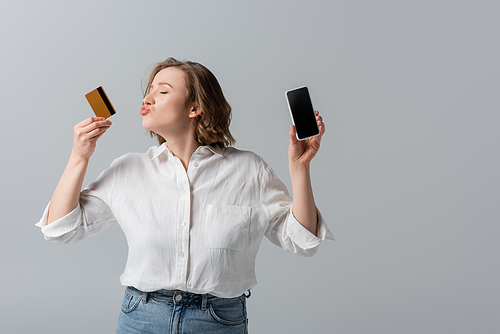 plus size woman holding credit card and kissing smartphone isolated on grey