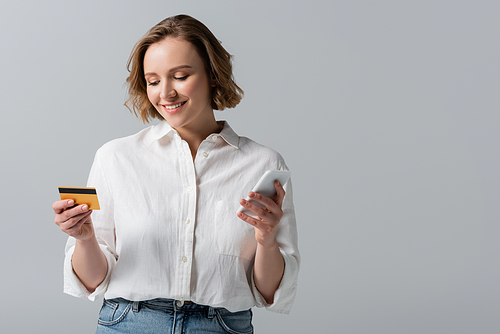 cheerful plus size woman holding credit card and smartphone isolated on grey