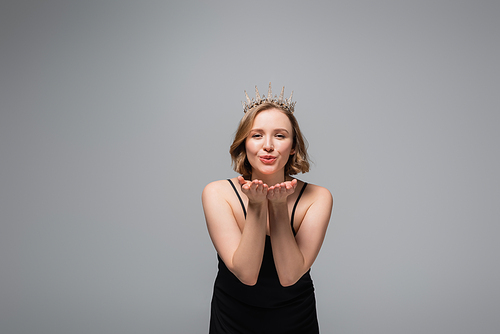 happy plus size woman in black slip dress and crown sending air kiss isolated on grey