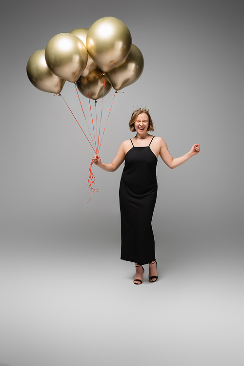 full length of excited plus size woman in black slip dress and crown holding golden balloons on grey