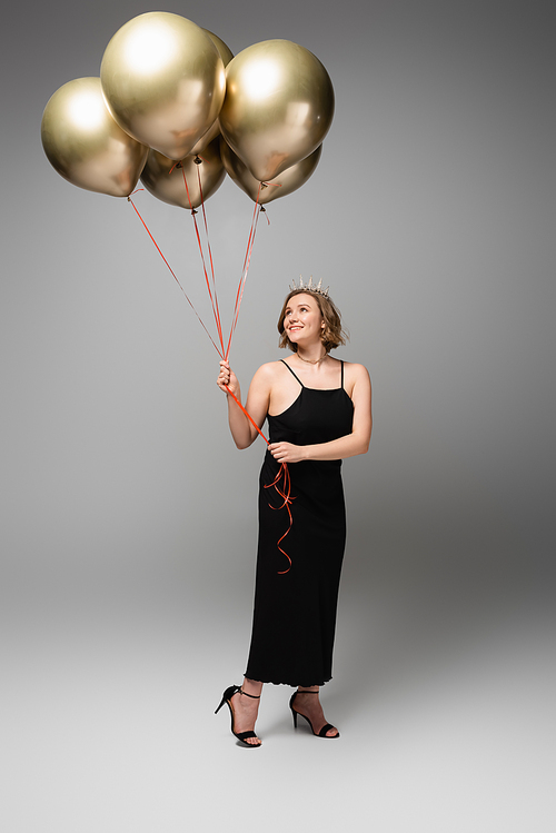 full length of smiling plus size woman in black slip dress and crown holding golden balloons on grey