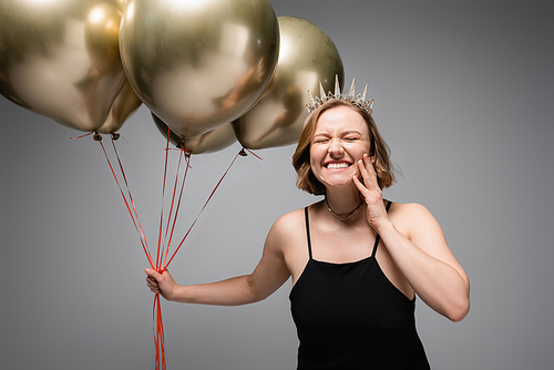 happy plus size woman in black slip dress and crown holding golden balloons on grey