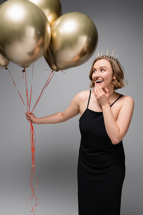 excited plus size woman in black slip dress and crown holding golden balloons on grey