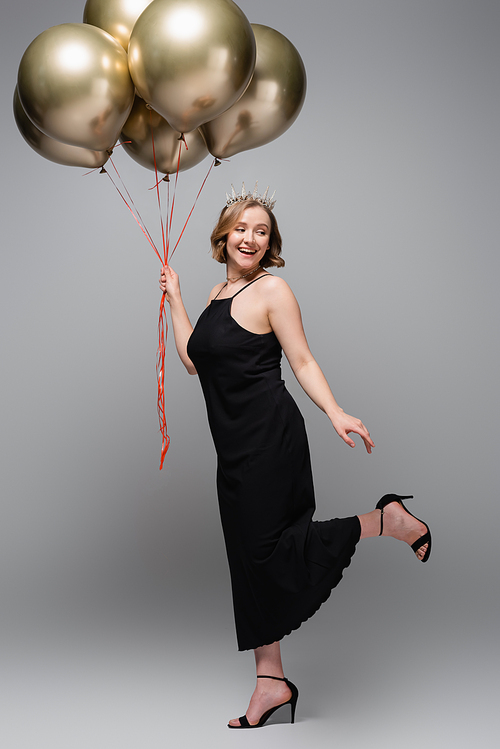 full length of joyful plus size woman in black slip dress and crown holding golden balloons on grey