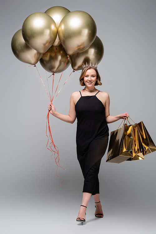 full length of happy plus size woman in black slip dress and crown holding golden balloons and shopping bags on grey