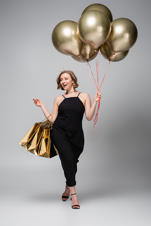 full length of happy plus size woman in slip dress and crown holding golden balloons and shopping bags on grey