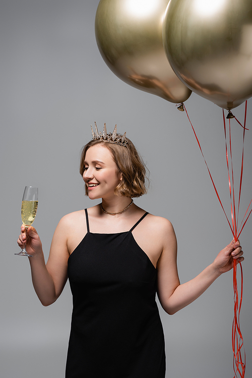 happy plus size woman in black slip dress and crown holding golden balloons and champagne isolated on grey