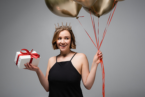 happy plus size woman in slip dress and crown holding golden balloons and present isolated on grey