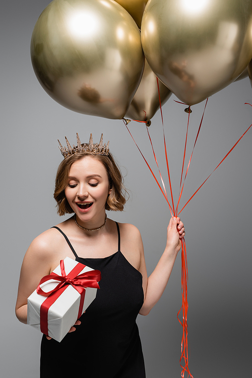 amazed plus size woman in slip dress and crown holding golden balloons and present isolated on grey