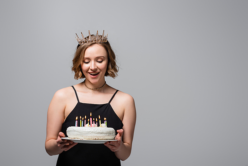 happy plus size woman in slip dress and crown holding birthday cake isolated on grey