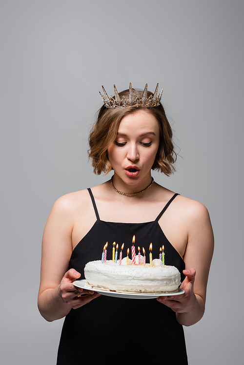 surprised plus size woman in slip dress and crown holding birthday cake isolated on grey