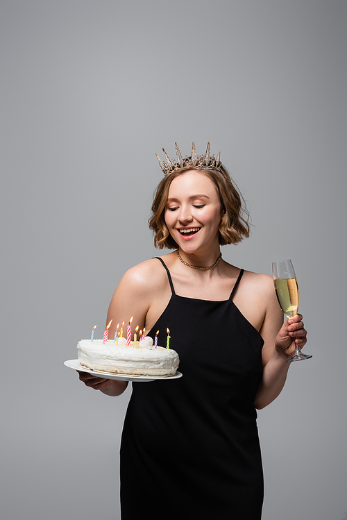 happy plus size woman in slip dress and crown holding birthday cake and glass of champagne isolated on grey