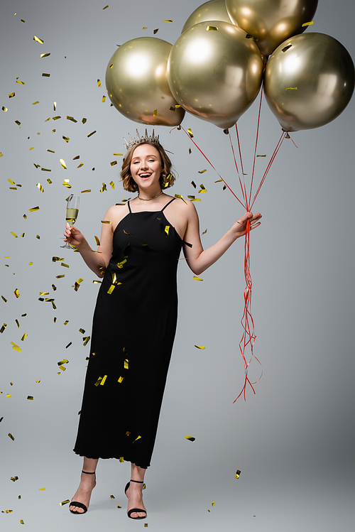 full length of happy plus size woman in slip dress and crown holding balloons and glass of champagne near confetti on grey