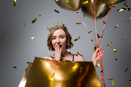 surprised plus size woman in crown holding balloons and shopping bags near confetti on grey