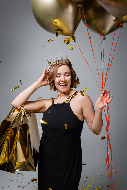 happy plus size woman in slip dress and crown holding golden balloons and shopping bags near confetti on grey