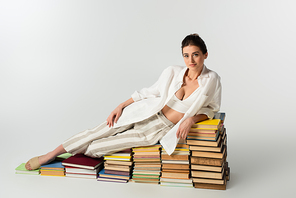 full length of brunette young woman in sandals lying on pile of books on white