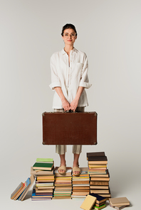 full length of pleased young woman in glasses holding suitcase while standing on pile of books isolated on white
