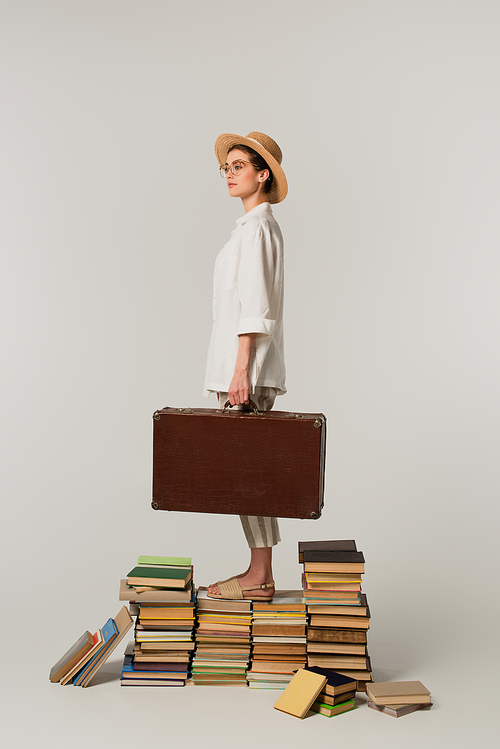 full length of young woman in straw hat and glasses holding suitcase while standing on pile of books isolated on white