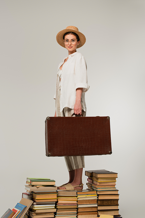 full length of pleased woman in straw hat and glasses holding baggage while standing on pile of books isolated on white