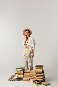 pleased young woman in sun hat standing near pile of books isolated on white