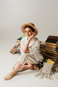 full length of smiling young woman in sun hat sitting near pile of books on white