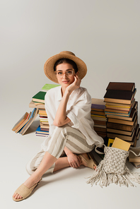 full length of pleased young woman in sun hat and glasses sitting near pile of books on white