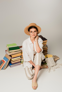 full length of pleased woman in sun hat and glasses sitting near pile of books on white