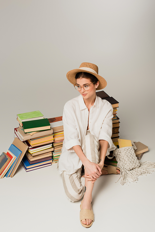 young woman in straw hat and glasses sitting near pile of books on white