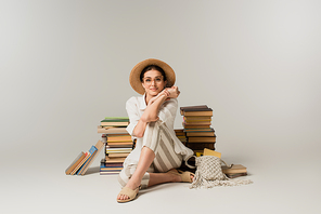 full length of happy young woman in sun hat and glasses sitting near pile of books on white
