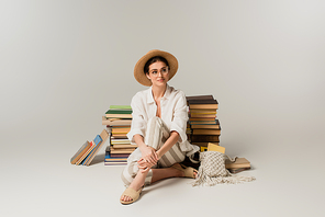full length of happy young woman in straw hat and glasses sitting near stack of books on white