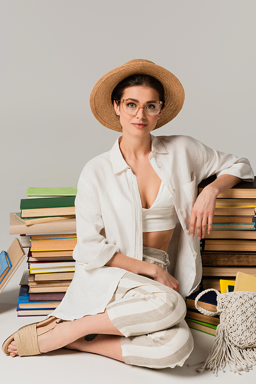 pleased woman in straw hat and glasses sitting near stack of books on white