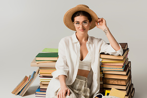 pleased and tattooed woman in straw hat and glasses sitting near pile of books on white