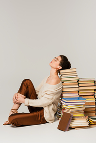 full length of young woman in glasses and sweater leaning on stack of books on white