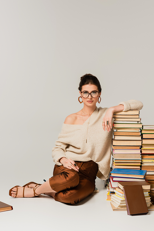 full length of young woman in glasses and sweater leaning on pile of books on white