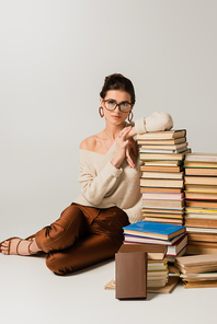 full length of young woman in glasses and earrings leaning on stack of books on white