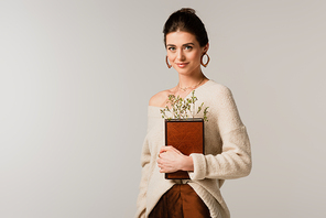 pleased young woman holding book with wildflowers isolated on grey