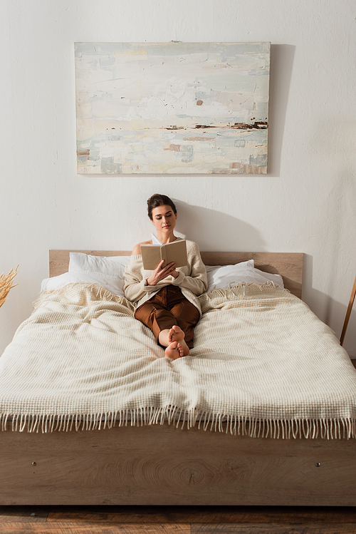 barefoot young woman reading book while resting on bed at home