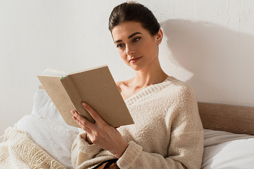 young woman reading book while resting on bed at home