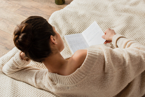 back view of young brunette woman in sweater reading book while resting on bed at home