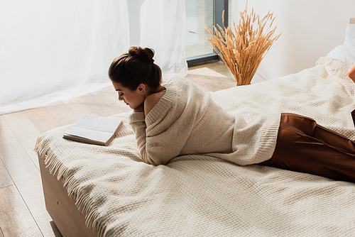 young woman reading book while resting on bed