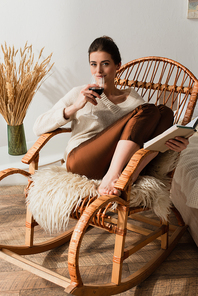 young woman sitting in wicker rocking chair with book and drinking red wine