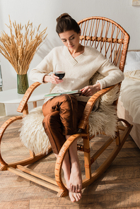 young woman sitting in wicker rocking chair with book and glass of red wine
